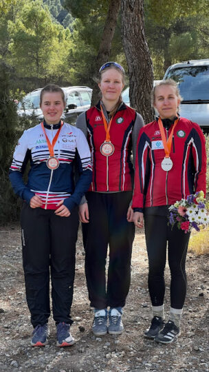 Tre av kvinnliga idrottare är uppställda jämsides, den första i vitt och blått och de andra i rött. De har alla en medalj runt halsen och Amy har en blombukett i handen.